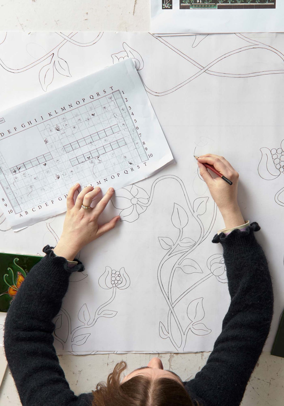 Top-down view of a person drawing floral line art on a large white sheet of paper, with a printed alphabet/design reference sheet nearby on a light neutral surface.