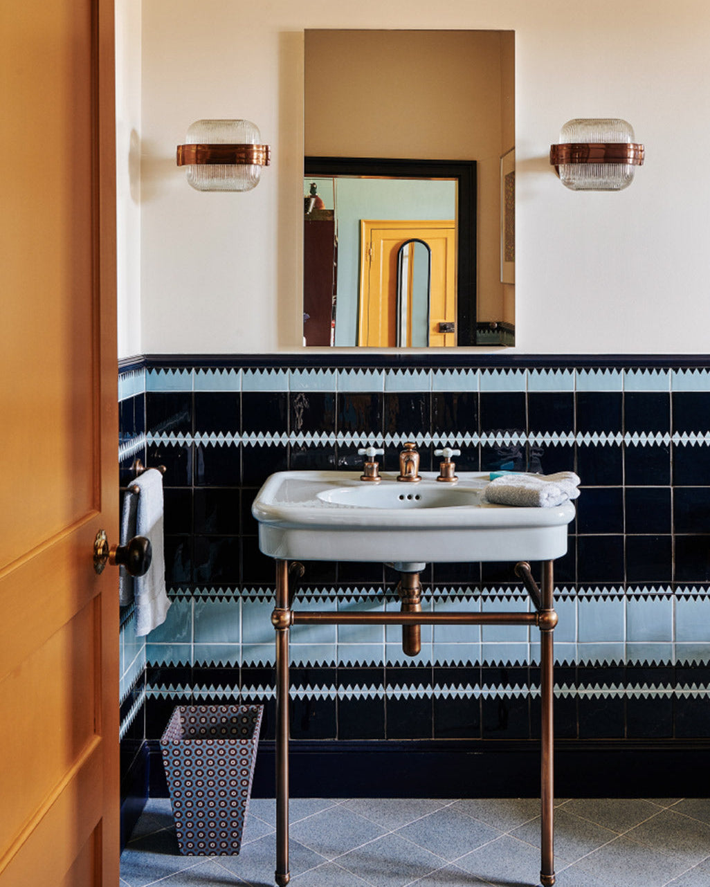 A vintage-style bathroom vanity with a white ceramic pedestal sink, brass faucet and legs, set against a wall with black and white patterned tile. A rectangular mirror hangs above, flanked by two wall sconces, and a wooden door is visible on the left.