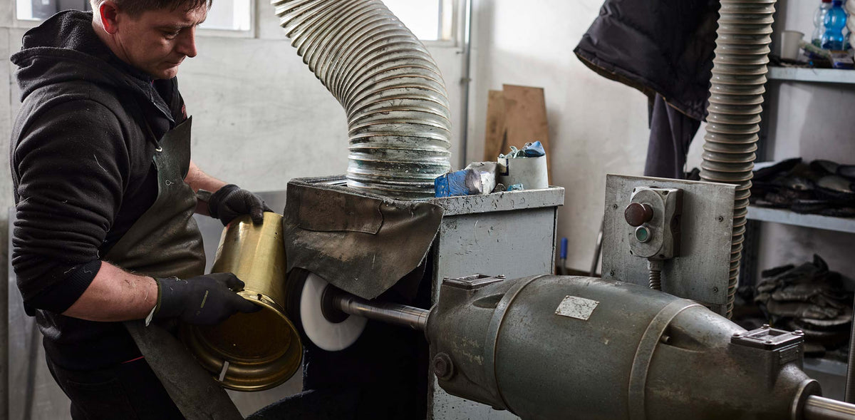 A man wearing a black hoodie and apron uses a metal grinder to shape a gold-colored metal part in an industrial workshop setting.