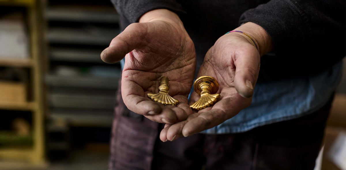 A close-up of a person's hands holding two small gold-colored decorative hardware pieces, likely cabinet knobs or pulls, against a blurred workshop background.