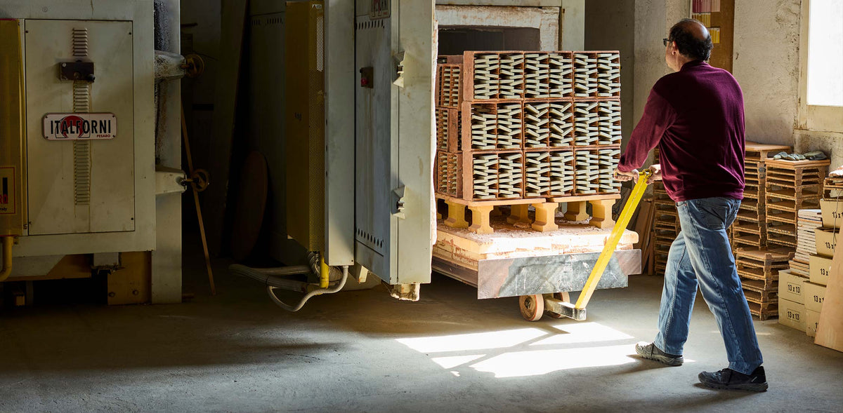 A worker in a warehouse pushes a pallet loaded with stacked rectangular items using a yellow handle, with a large machine and wooden pallets visible in the industrial setting.