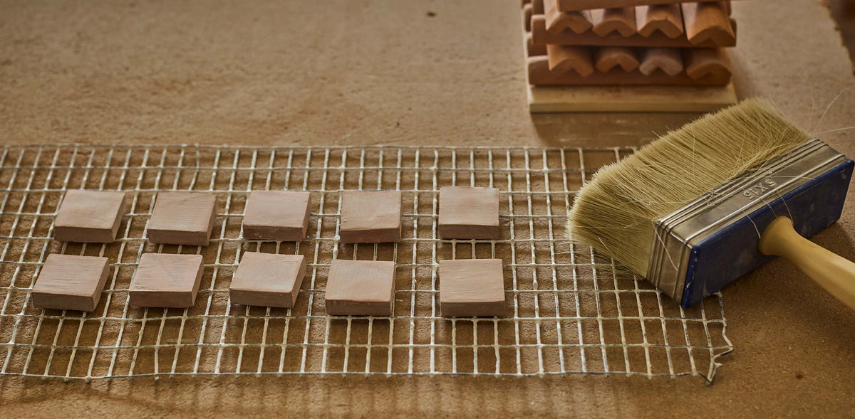 Small square wooden tiles arranged on a metal mesh tray, with a paintbrush resting on the right side and a stack of similar tiles in the background on a tan surface.