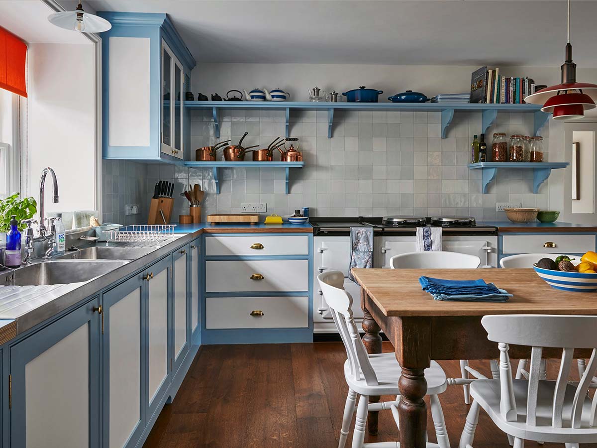 A bright kitchen interior with light blue and white cabinets, open blue shelves holding cookware and books, a tiled backsplash, dark wood flooring, and a wooden dining table with white chairs in the foreground.
