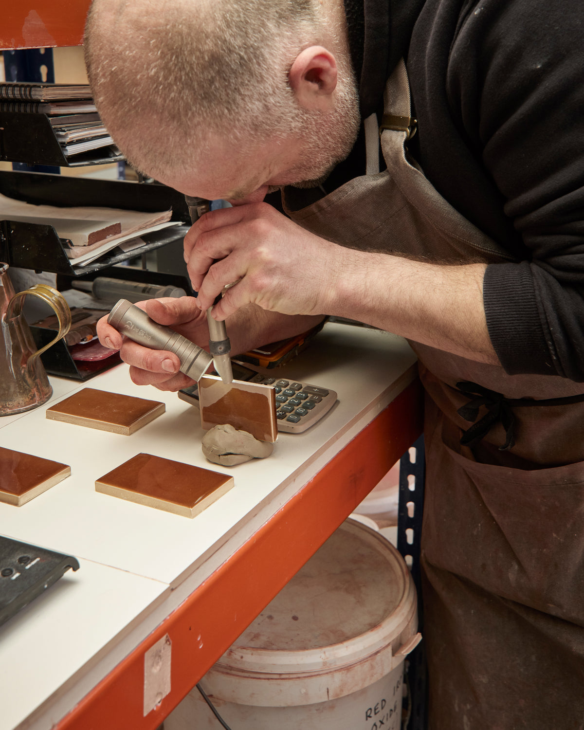 A craftsman wearing a brown apron leans over a workbench, using a handheld tool on a small wooden sample while other wood blocks and tools are arranged on the white table.